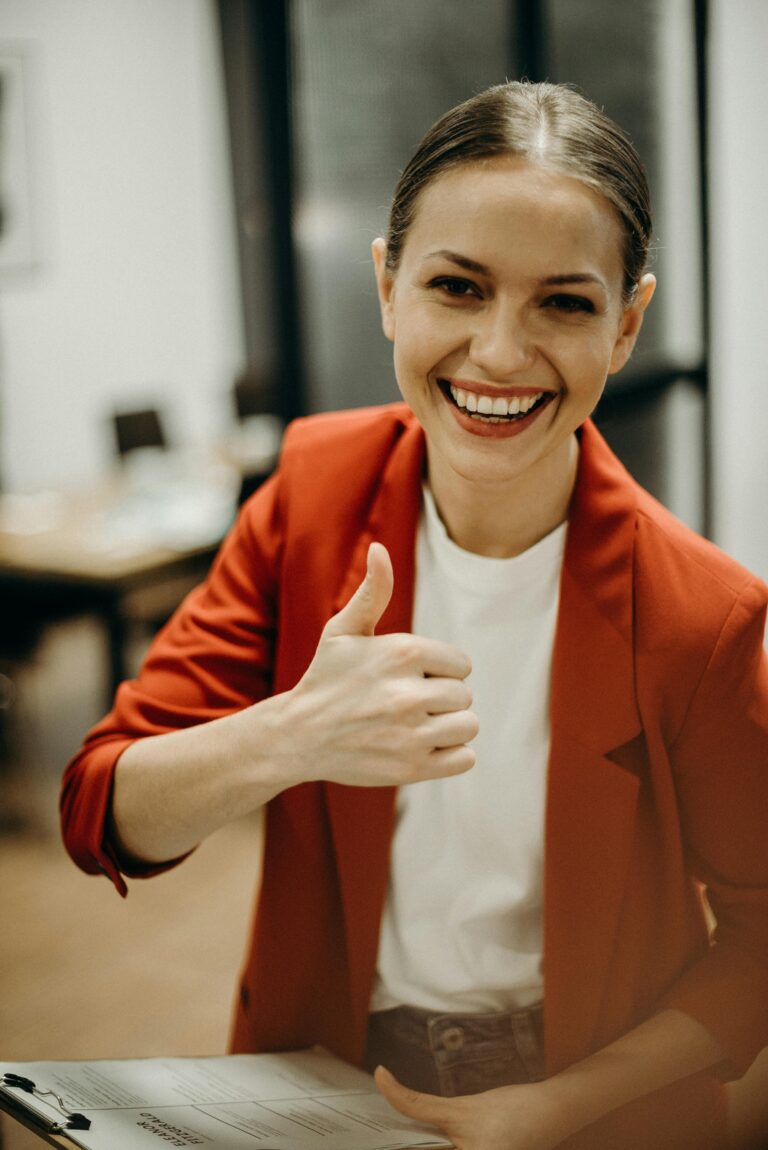 Confident businesswoman in a red blazer gives a thumbs up in a modern office setting.